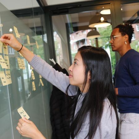 Two people are working collaboratively on a white board, using sticky notes to organize information