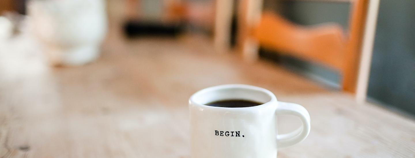 A cup of hot coffee in a white mug that reads: Begin. on a wooden table.