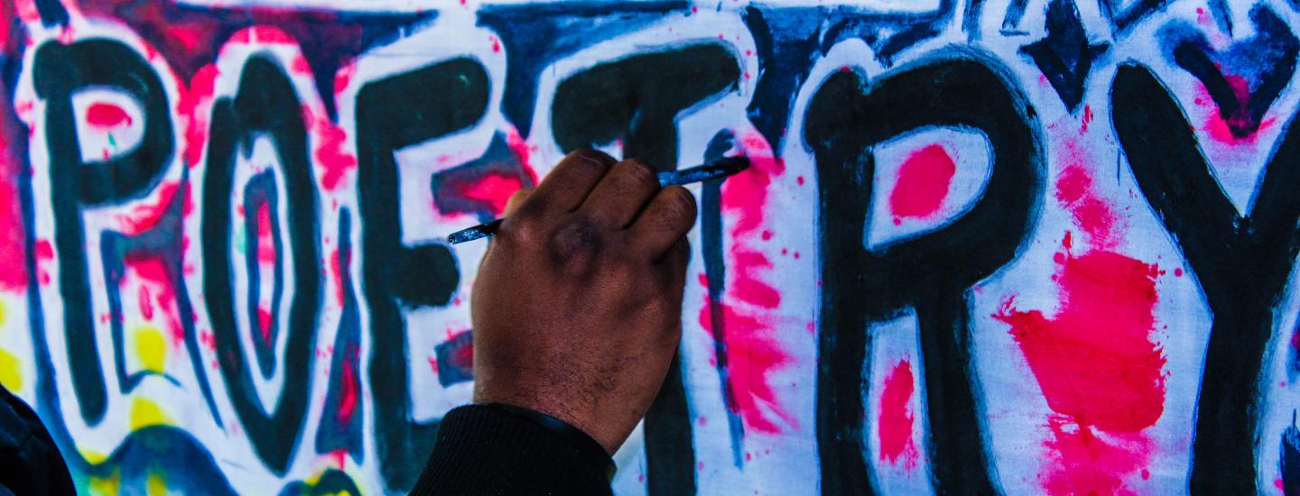A black man's hand is painting the word "poetry" on a wall, graffiti style