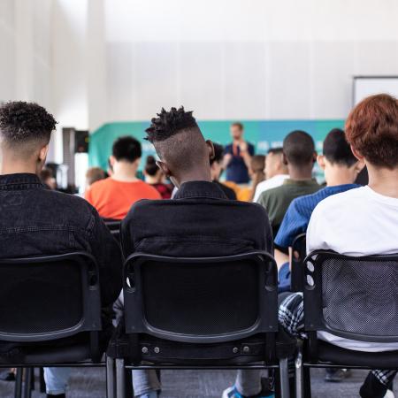 Students sit in a packed classroom viewed from behind. 